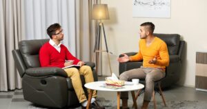 Two men sit in armchairs across from each other in a living room, one taking notes in a red sweater, the other gesturing in an orange sweater. -